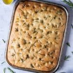 Loaf of sea salt and rosemary focaccia in a baking tin on a marble background with fresh rosemary and bowls of olive oil and sea salt.