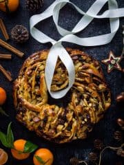 A top down shot of a braided bread wreath filled with fruit mince, marzipan and apple; surrounded by clementines, spices and Christmas decorations