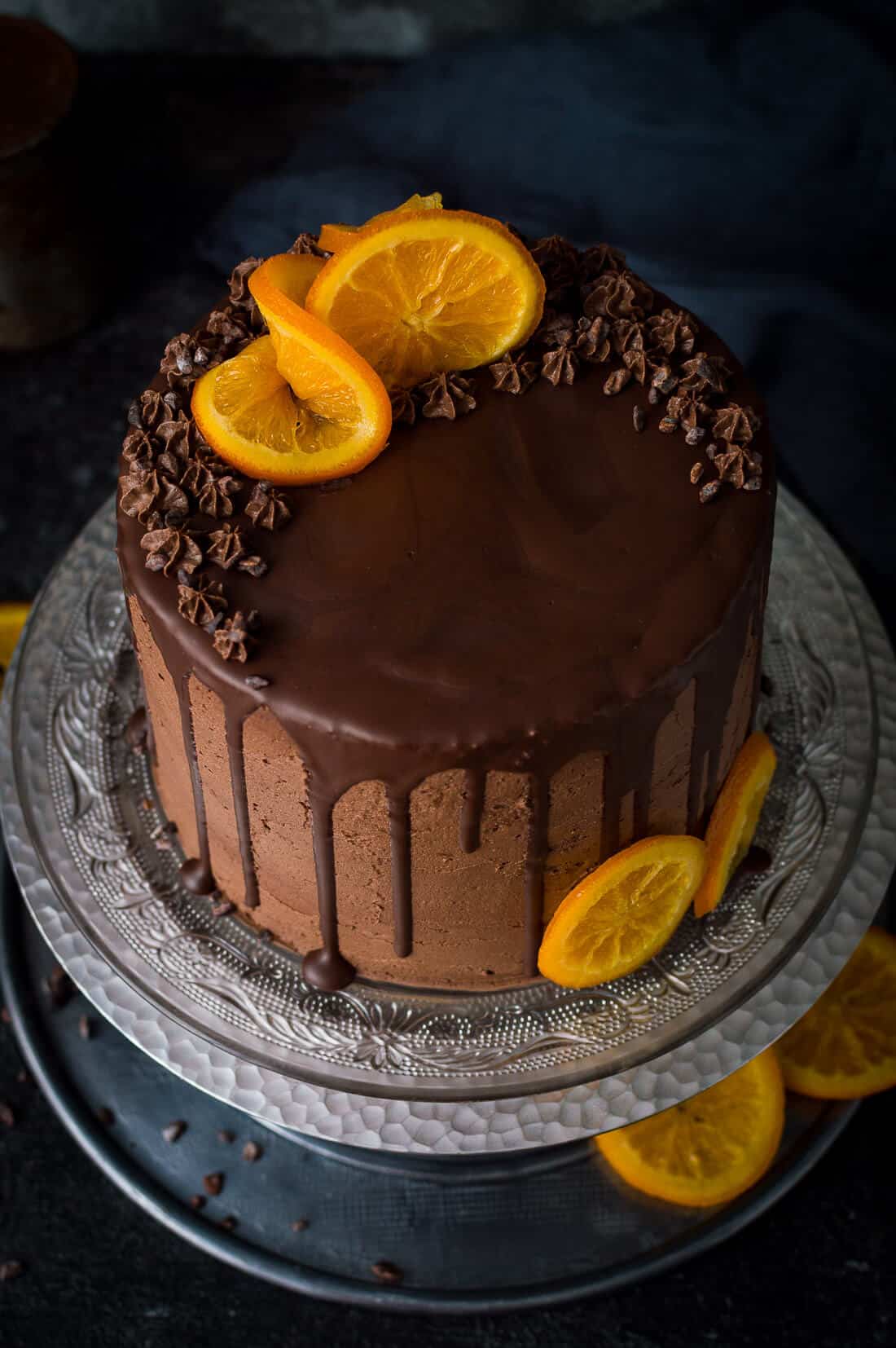 Angled photo of vegan orange and almond cake with chocolate orange vegan buttercream, chocolate drip and candied oranges on a glass plate and silver cake stand.