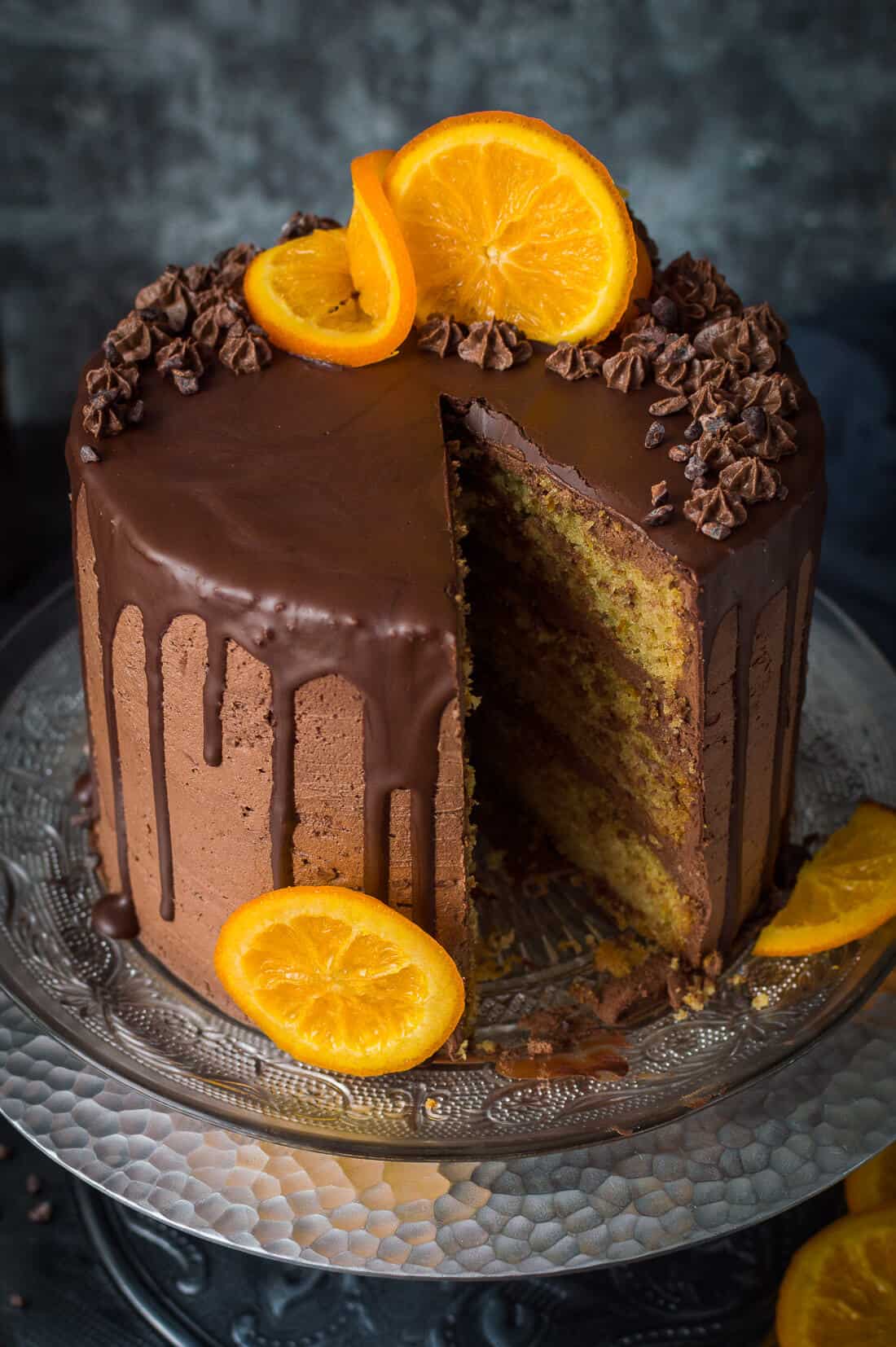 Vegan orange and almond layer cake with chocolate buttercream and candied oranges on a silver cake stand with a slice taken out.