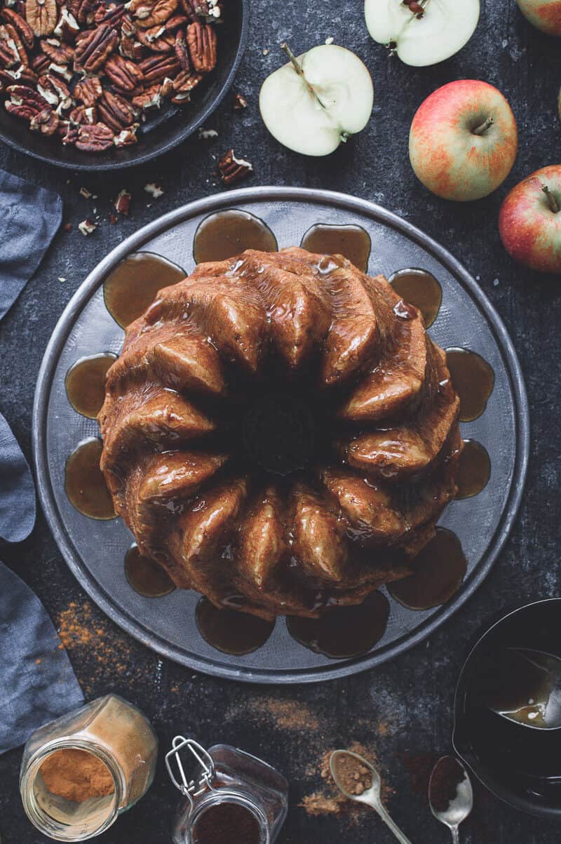 Top down shot of vegan apple bundt cake on a metal plate on a black background with pecans, apples and spices.