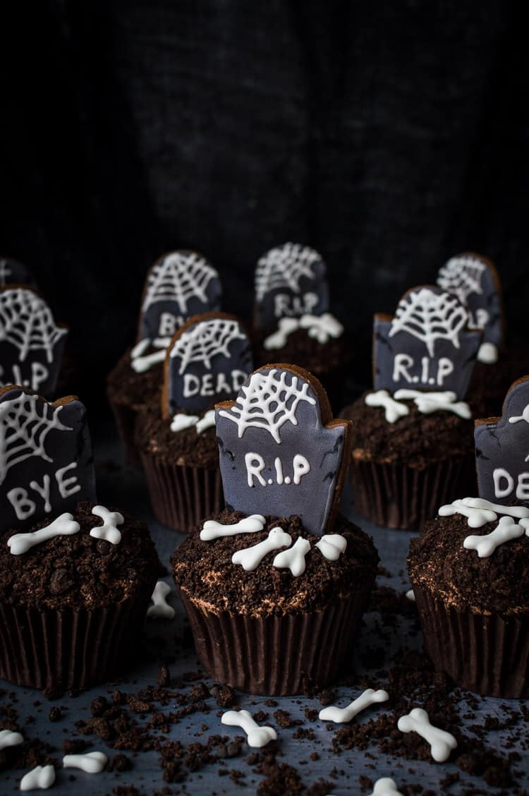 Vegan chocolate cupcakes topped with crushed Oreos, gingerbread tombstones and vegan royal icing bones on a dark background.