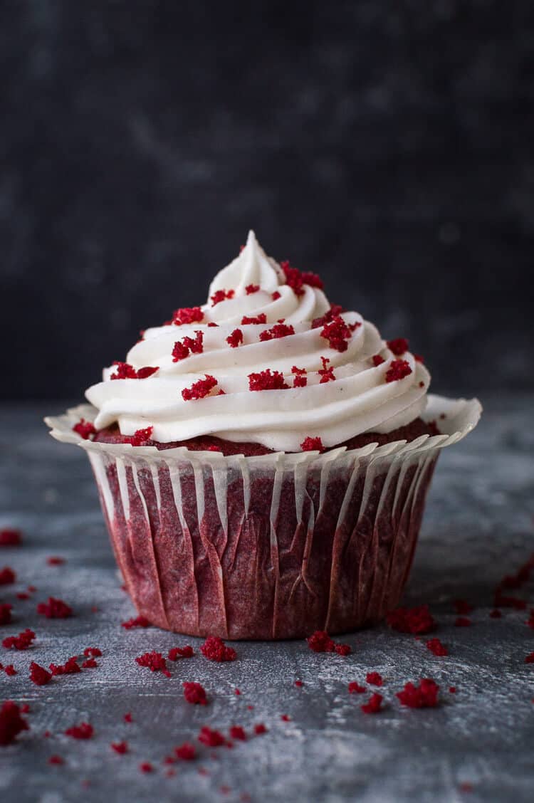 A vegan red velvet cupcake with cream cheese frosting and cake sprinkles on a grey background.