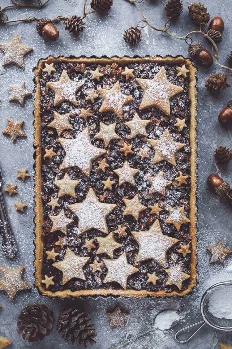 Starry mince pie tart dusted with icing sugar on a grey background surrounded by cinnamon shortcrust stars, pine cones and Christmas decorations.