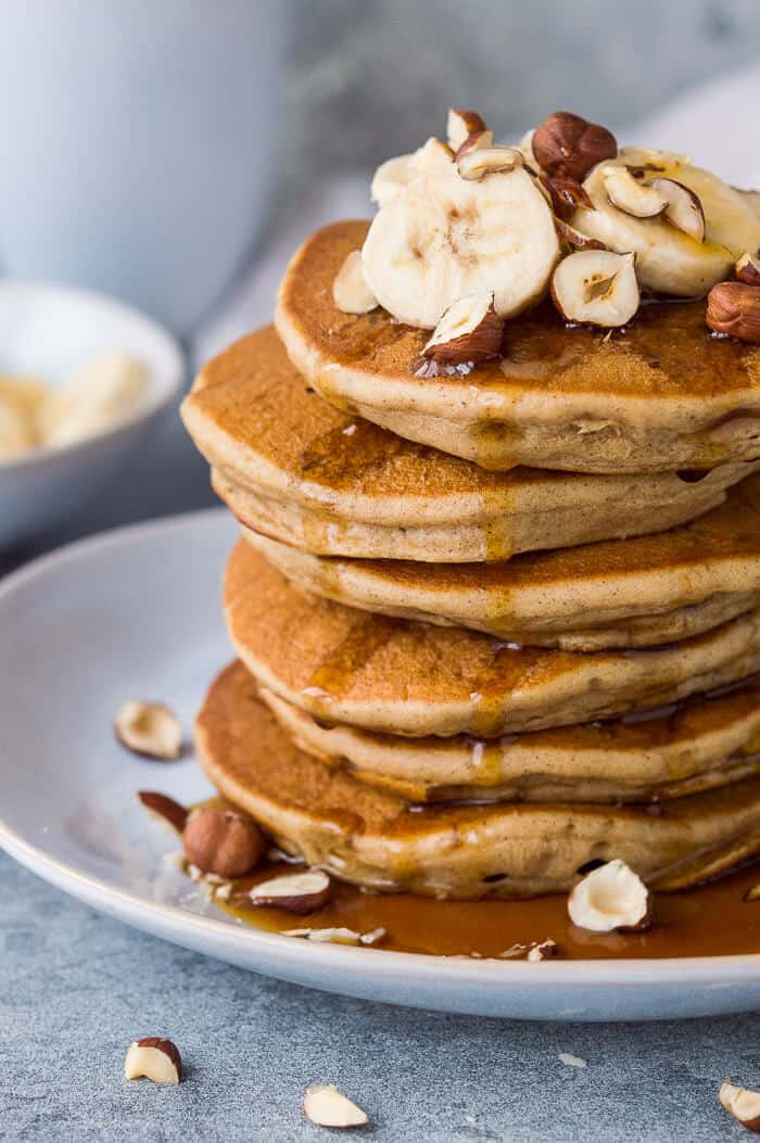 Close up of a stack of fluffy vegan banana pancakes topped with sliced banana, hazelnuts and maple syrup.