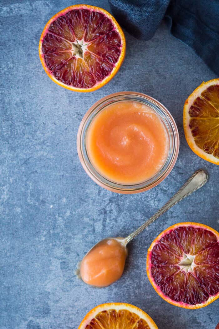 Vegan blood orange curd in a glass jar on a blue/grey background with sliced blood oranges and a teaspoon. 