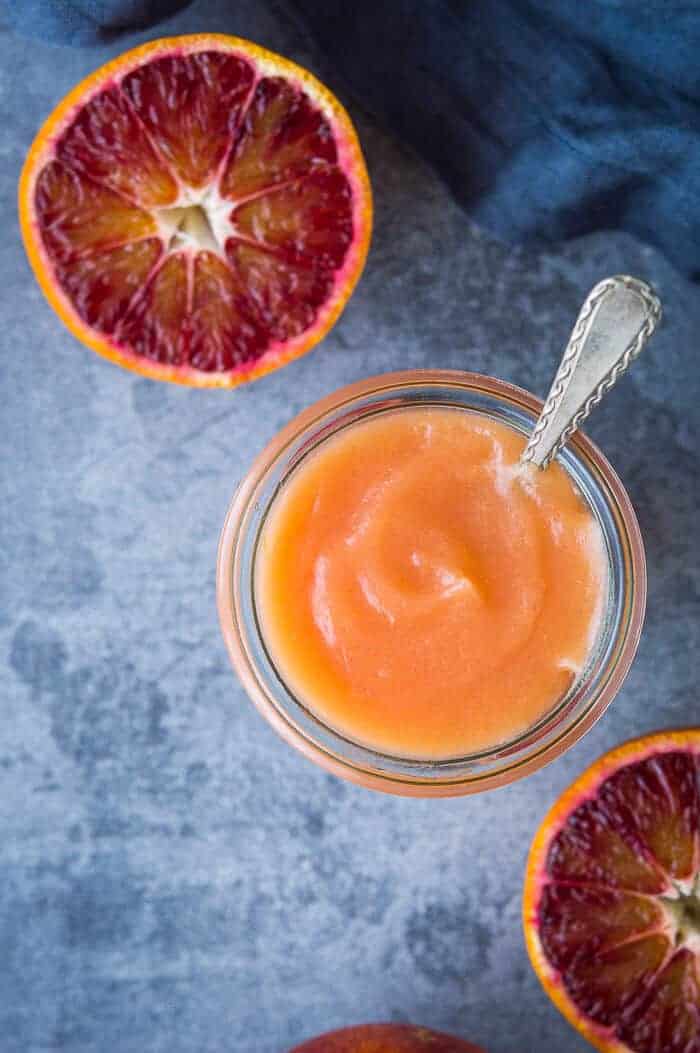 close up of a jar of vegan blood orange curd with a spoon in it and sliced blood oranges.