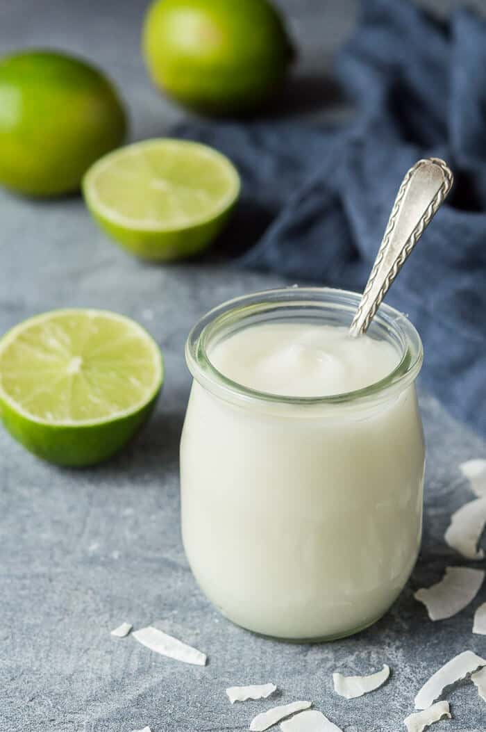 A small glass jar of coconut lime curd on a grey background with limes and shredded coconut.