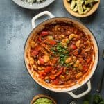 A pan of vegan vegetable, lentil and butter bean chilli with a bowl of coriander rice, roasted courgettes and guacamole on a grey background.