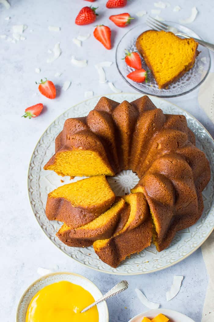 eggless mango bundt cake on a white plate with a slice of cake, strawberries, mango and coconut.