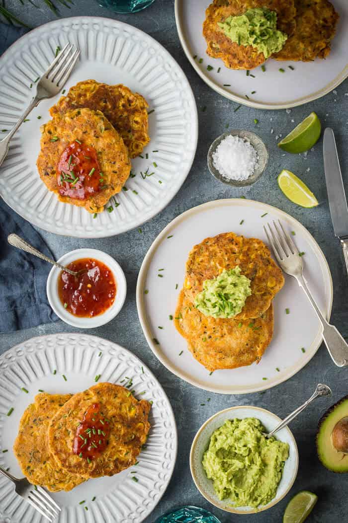 Four white plates of vegan corn fritters with guacamole and chilli sauce on a grey background.