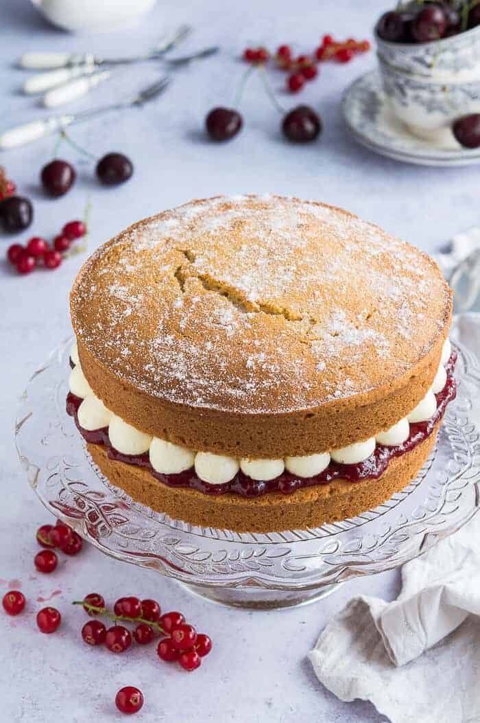 Vegan Victoria sponge cake filled with jam and buttercream on a glass cake stand on a grey background with cherries and redcurrants.