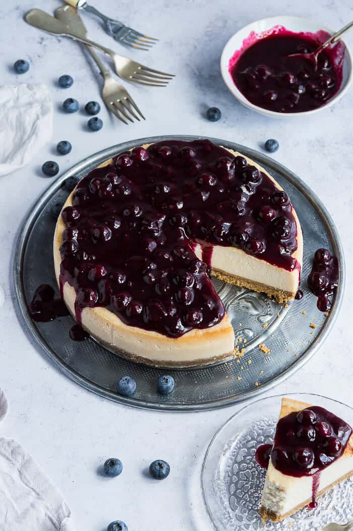 vegan baked cheesecake on a metal platter on a grey background with blueberries, blueberry compote and forks.