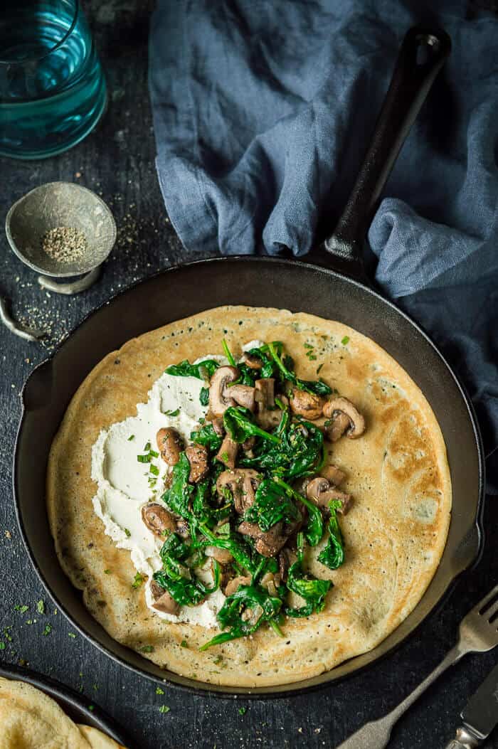 Vegan spinach, mushroom cheese crepe in a cast iron pan on a black background with a grey cloth and pewter dish of pepper.