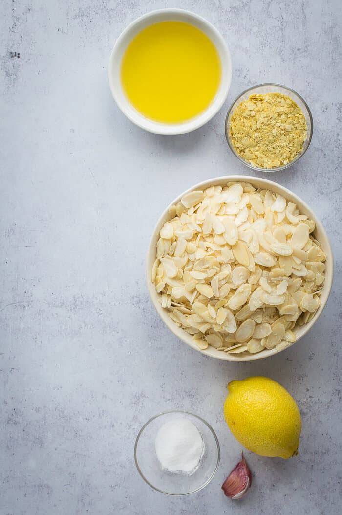 The ingredients needed to make vegan almond cream cheese laid out on a grey background - bowls of flaked almonds, olive oil, nutritional yeast, salt, lemon and garlic.