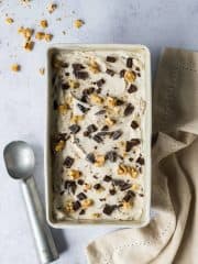 vegan chunky monkey ice cream in a loaf pan with an ice cream scoop and a beige cloth on a grey background.