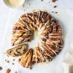 Maple pecan bread wreath with two slices cut out on a sheet of baking parchment with a bowl of glaze, pecan nuts and mini bottles of maple syrup.