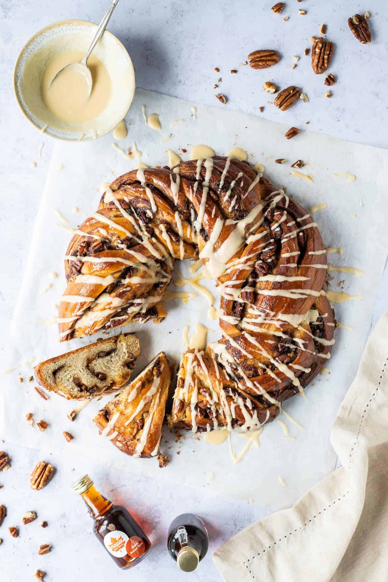 Maple pecan bread wreath with two slices cut out on a sheet of baking parchment with a bowl of glaze, pecan nuts and mini bottles of maple syrup.