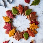 Autumn leaf maple cookie wreath on a white background with coloured leaf cookies, cookie cutters and bottles of maple syrup.