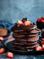 A stack of vegan chocolate pancakes on a black plate topped with chocolate sauce and fresh berries.