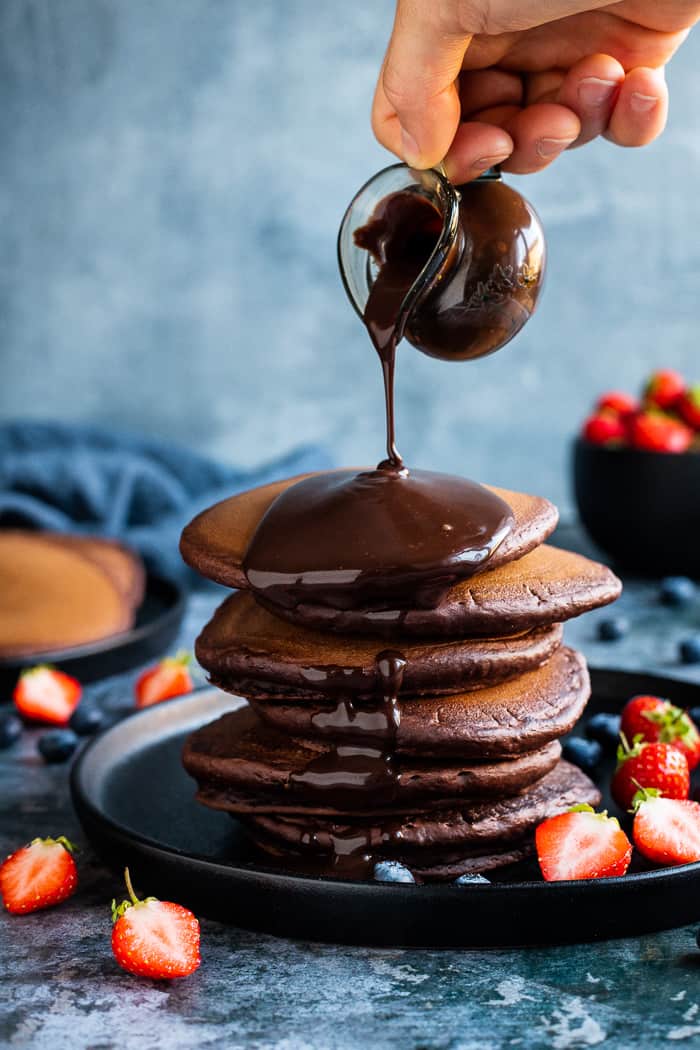 Chocolate sauce being poured onto a stack of chocolate pancakes from a small glass jug.