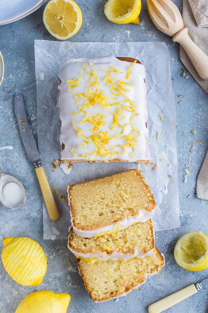Sliced vegan lemon drizzle cake on a grey background surrounded by whole and sliced lemons, a lemon juicer, a knife and plates.