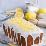Vegan lemon drizzle cake with lemon glaze and candied lemon zest on a sheet of baking parchment with lemons and crockery in the background.