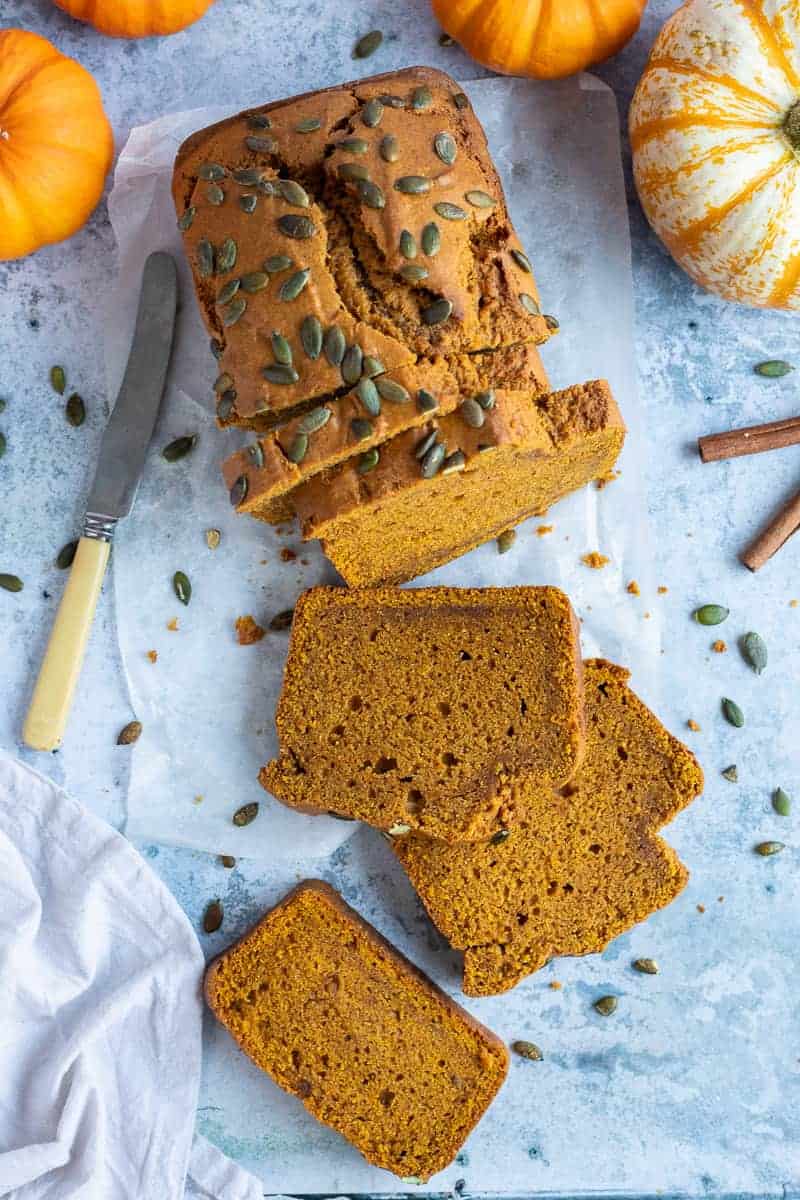a loaf of vegan pumpkin bread topped with pumpkin seeds on a sheet of baking parchment on a blue background with mini pumpkins, cinnamon sticks, a grey cloth and a knife.