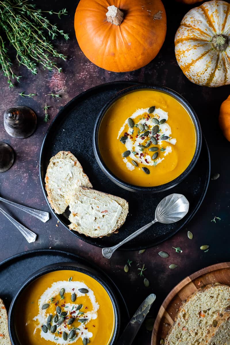 Two black bowls of vegan carrot and pumpkin soup on black plates on a dark background with mini pumpkins, bread and fresh thyme sprigs.