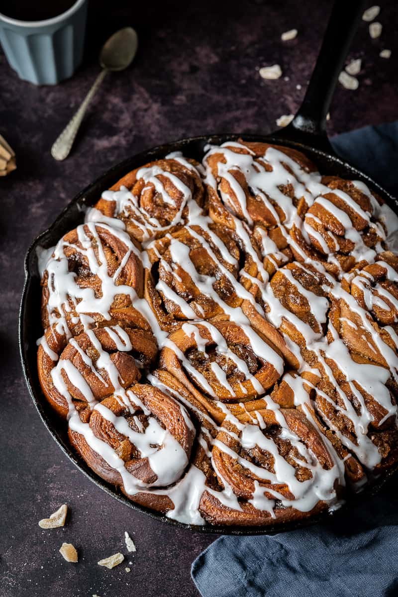 A skillet of ginger cinnamon rolls on a dark background with chopped candied ginger, a grey cup of espresso and a teaspoon.