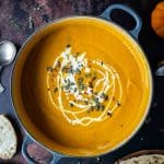 A panful of spiced carrot and pumpkin soup with a swirl of cream and pumpkin seeds on a dark background with mini pumpkins and slices of bread.