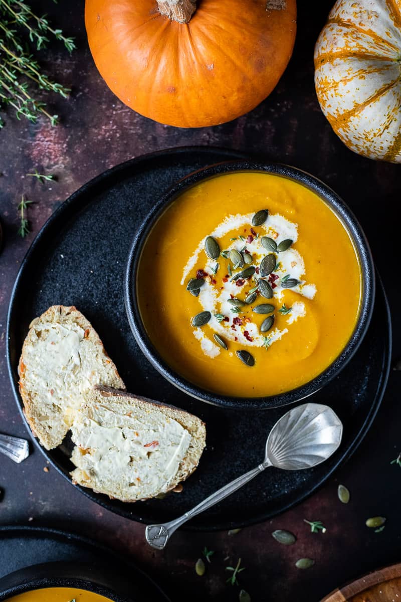 Close up of a black bowlful of spiced carrot and pumpkin soup on a black plate topped with a swirl of cream, pumpkin seeds and chilli flakes.