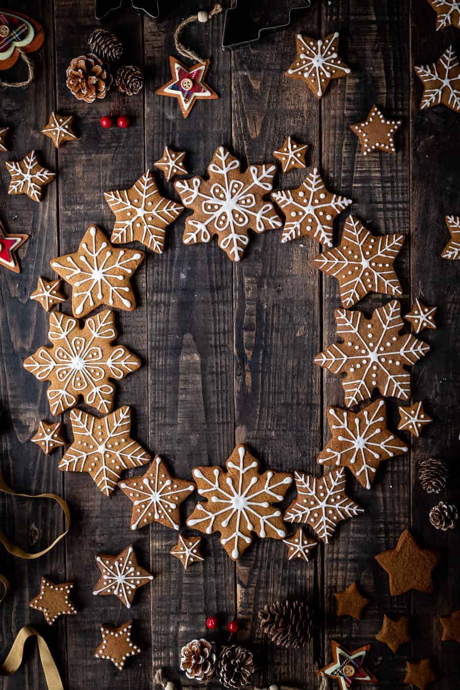 Vegan gingerbread snowflake cookies arranged in a wreath shape on a dark wood background with pine cones and Christmas decorations.