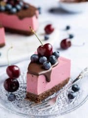 A slice of vegan chocolate cherry cheesecake topped with blueberries and a cherry on a glass plate with a forkful removed.