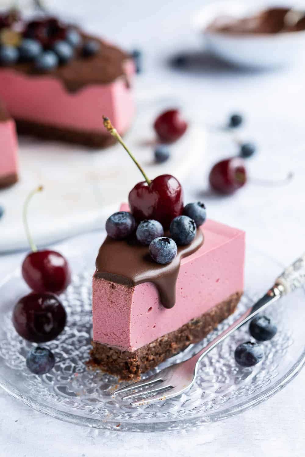 A slice of vegan chocolate cherry cheesecake topped with blueberries and a cherry on a glass plate with a forkful removed.