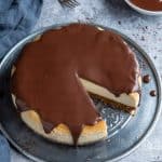 Cheesecake topped with chocolate ganache on a metal plate on a grey background with a bowl of ganache and three forks.