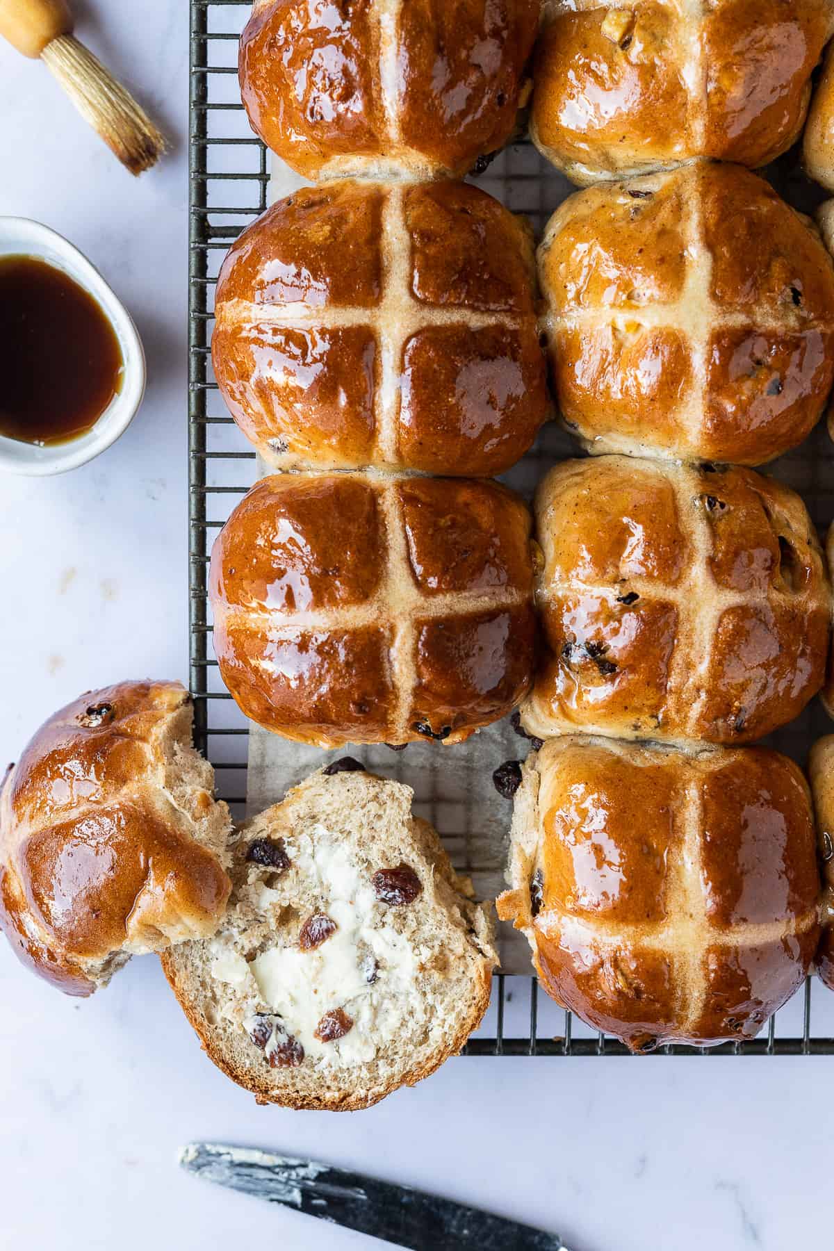 vegan hot cross buns on a wire rack, one split and buttered with a bowl of maple syrup and a brush.