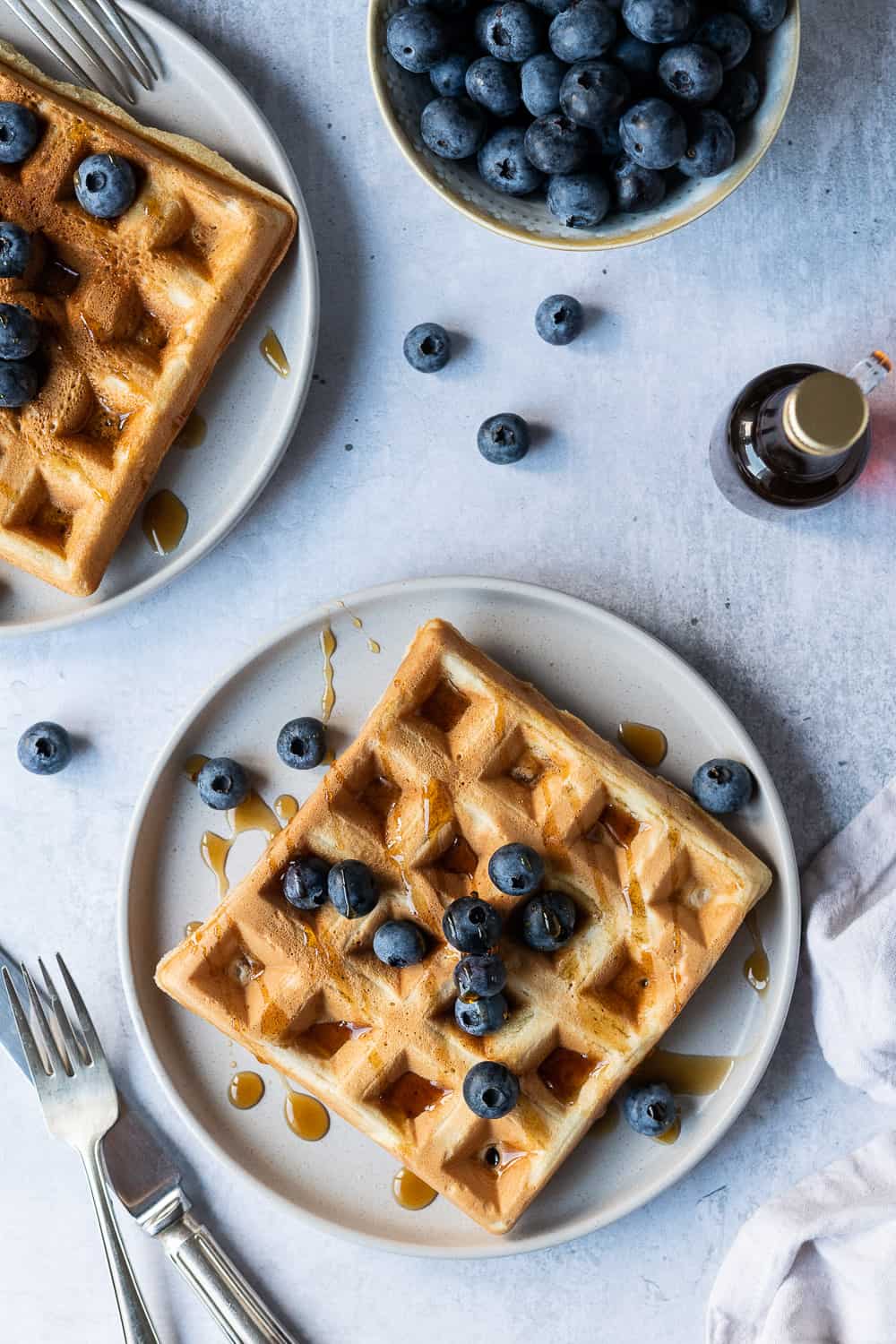 Two vegan waffles on grey plates with blueberries and maple syrup.