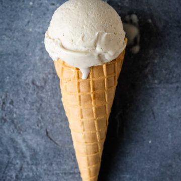 A waffle cone with a single scoop of vegan vanilla ice cream on a grey background.