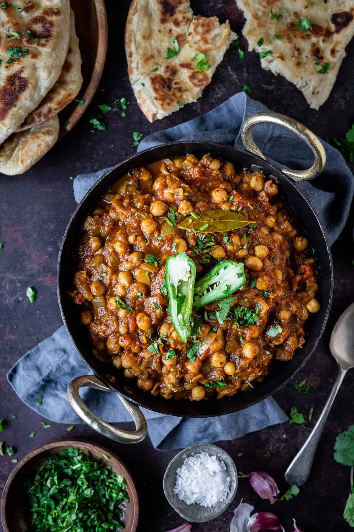 Chana masala in a black metal dish on a dark background with naan bread, fresh coriander, a pot of salt and a spoon.