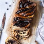 A loaf of vegan chocolate babka on a sheet of brown baking parchment on a grey background with a bowl of chocolate chips, a knife and a pastry brush.