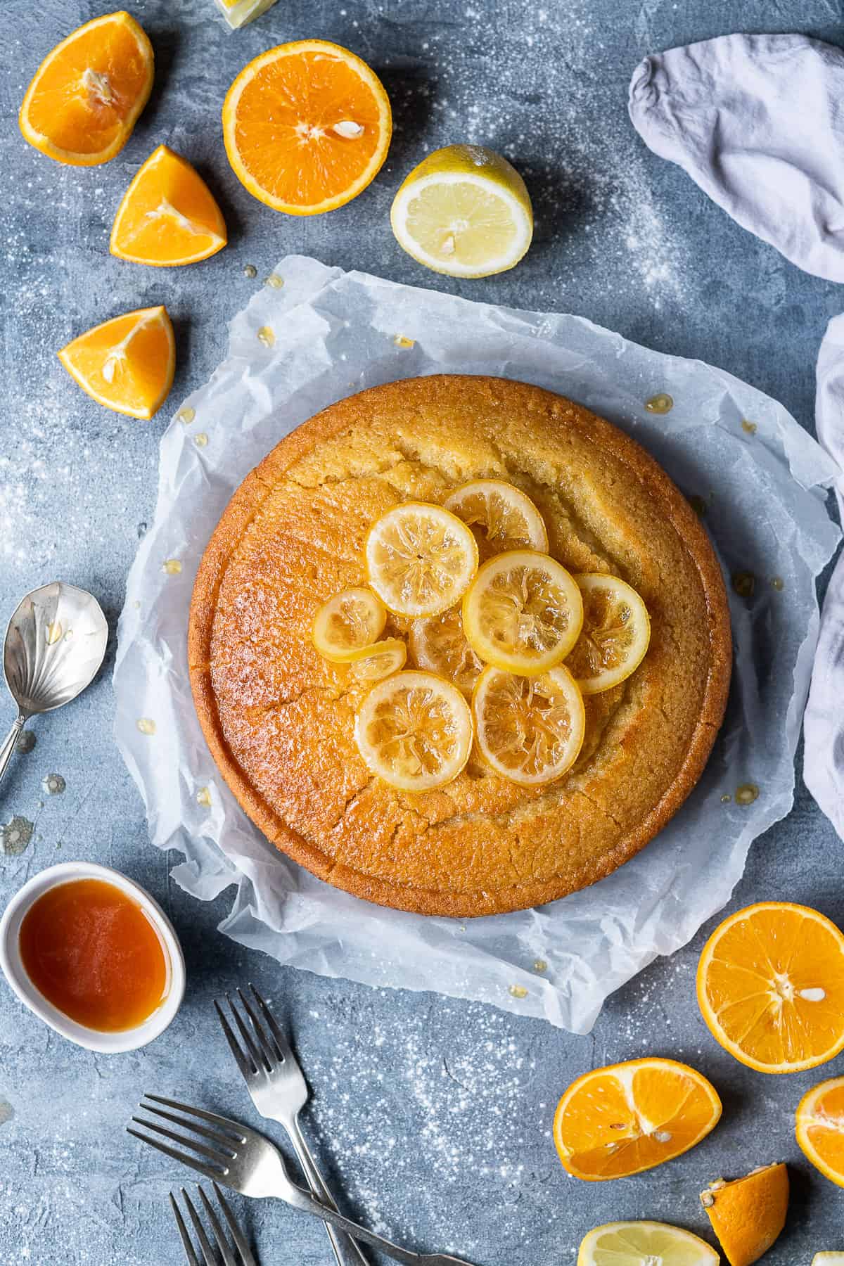 Vegan semolina cake topped with candied lemon slices on a sheet of baking parchment on a grey background with sliced oranges and lemons and three forks.