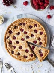 vegan raspberry Bakewell tart on a round marble board on a white background with a bowl of raspberries.