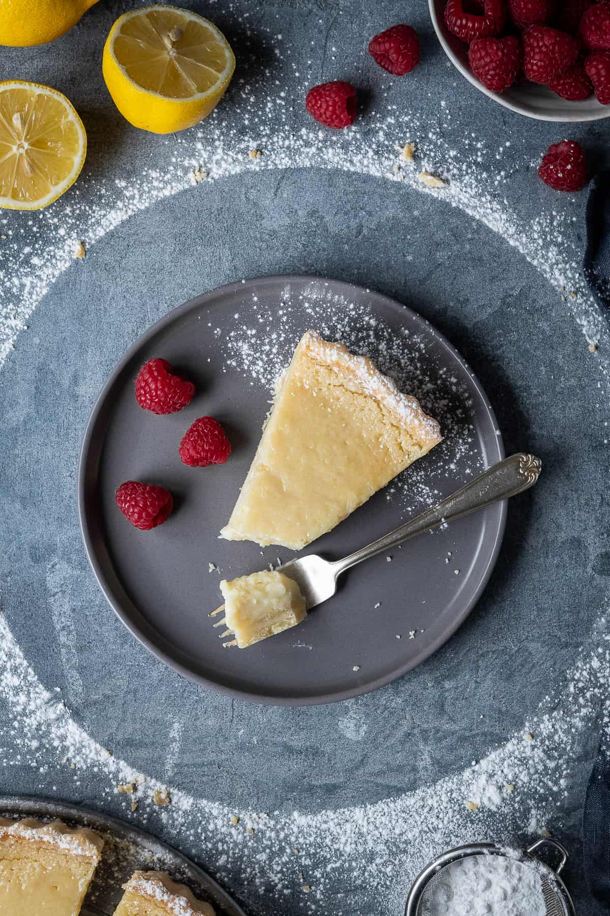 A slice of vegan lemon tart with raspberries and a fork on a grey plate