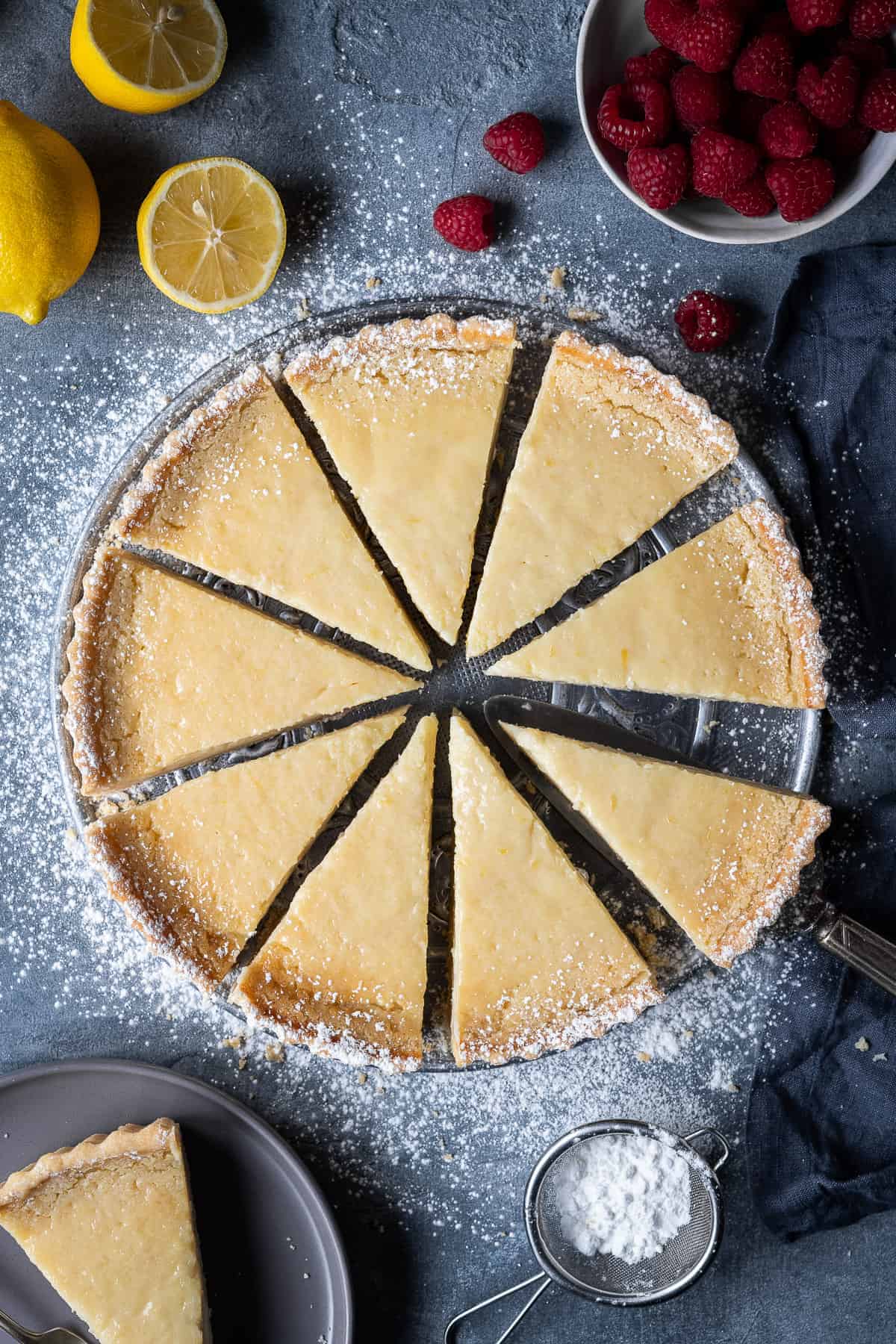 Sliced vegan lemon tart on a grey background with lemons and raspberries.