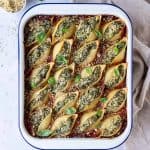Vegan spinach and ricotta stuffed pasta shells in a white enamel dish on a marble background with fresh basil, vegan parmesan and a cream cloth.