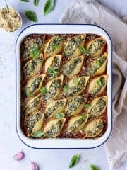 Vegan spinach and ricotta stuffed pasta shells in a white enamel dish on a marble background with fresh basil, vegan parmesan and a cream cloth.