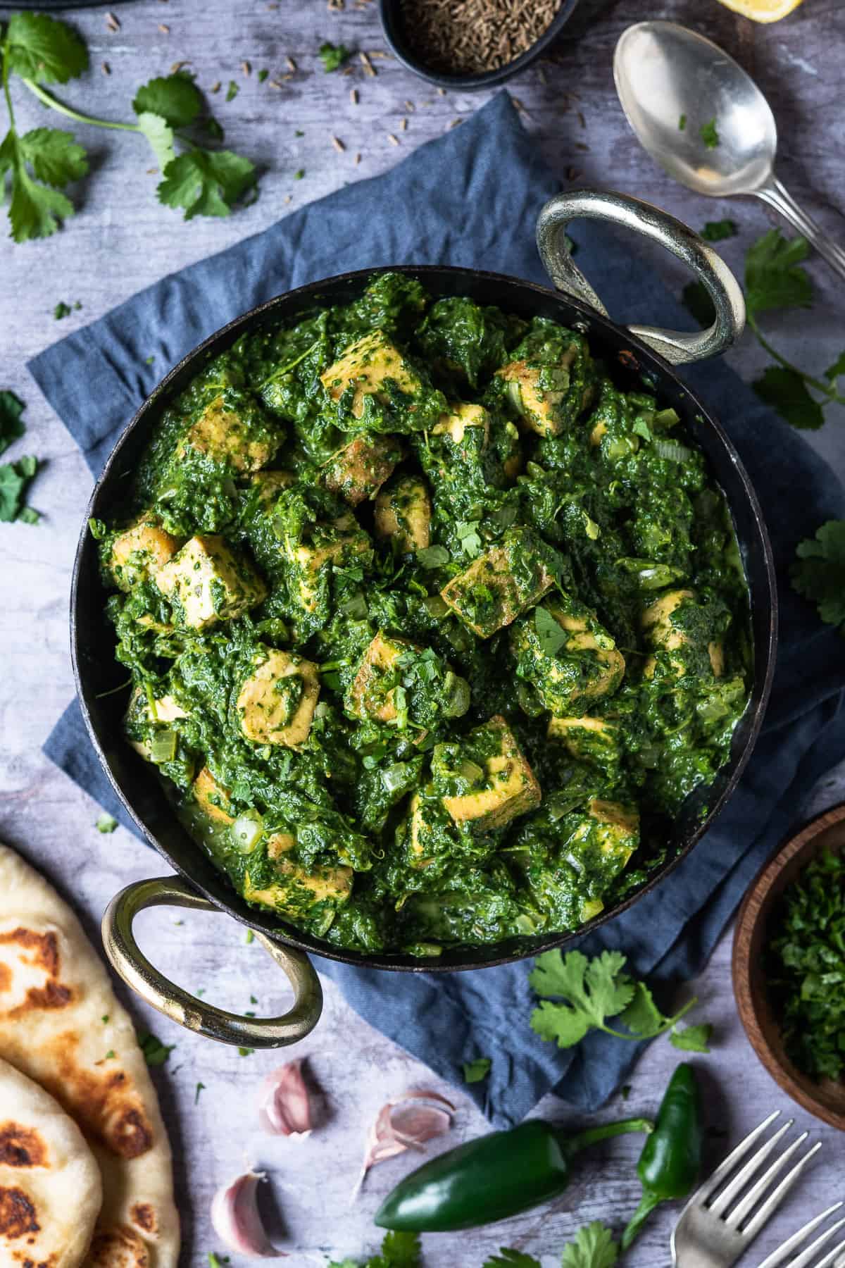 Vegan palak paneer in a kadhai pan on a grey surface surrounded by naan breads, coriander, green chillies and garlic cloves.