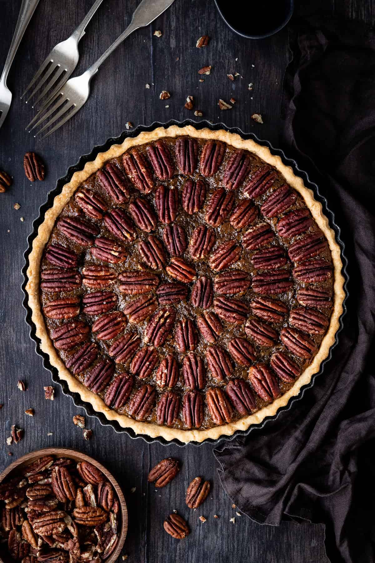 Vegan pecan pie on a wooden backdrop with a bowl of pecans, forks and a brown cloth.