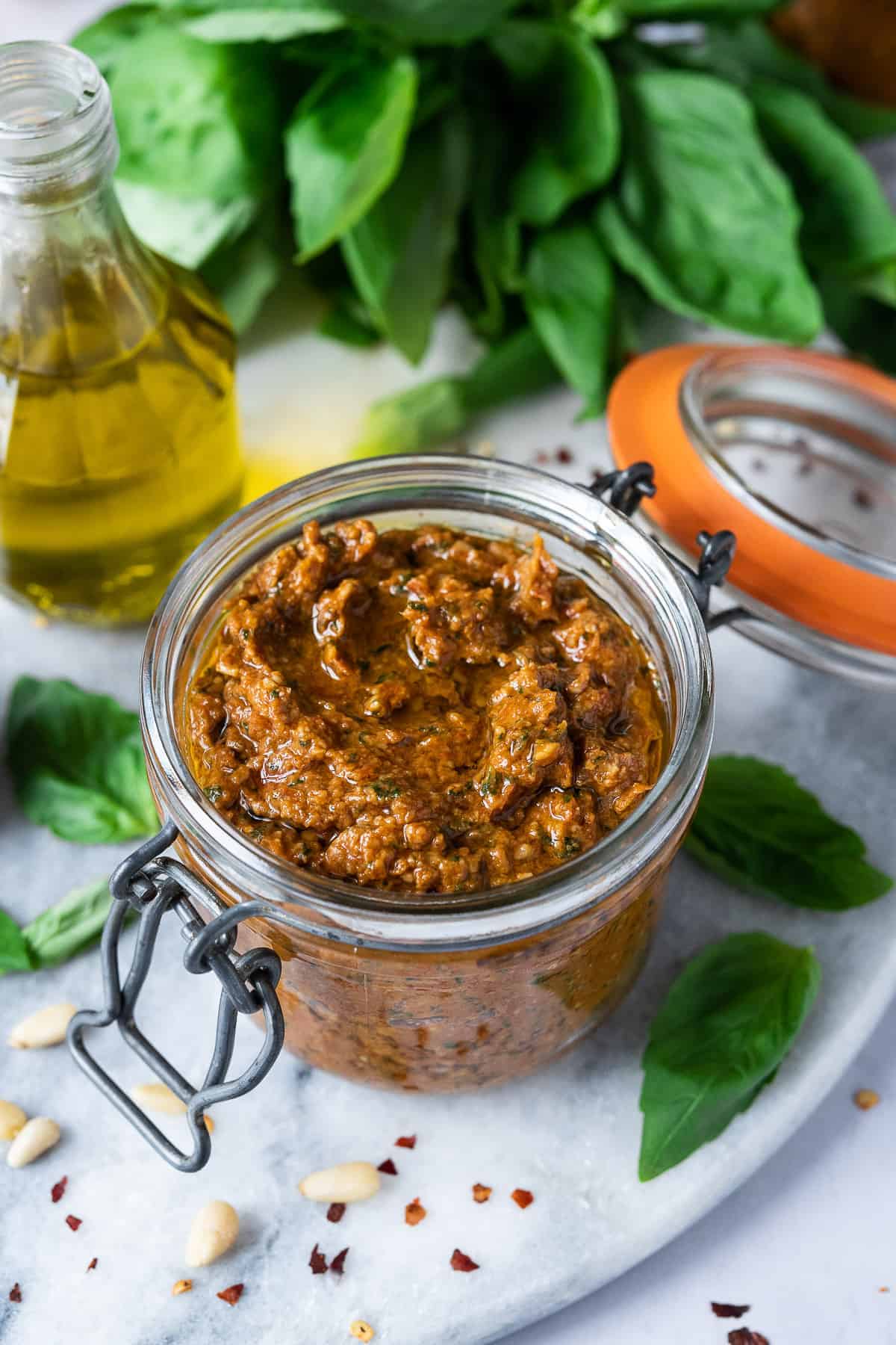 A jar of vegan sun dried tomato pesto on a marble board with fresh basil leaves and a bottle of olive oil.